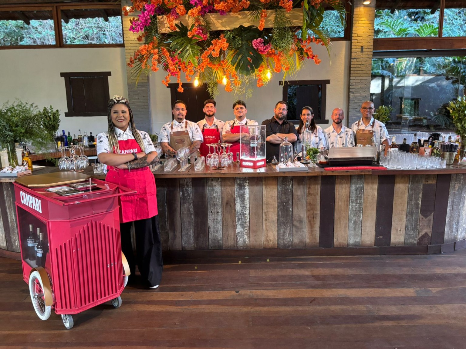 Registro da Alkimia em um evento: equipe reunida em um bar de madeira, com taças e flores tropicais
